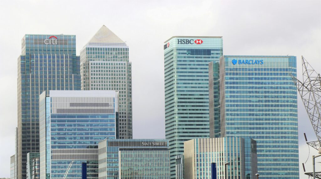 A cityscape featuring modern glass skyscrapers with logos of major financial institutions, including Citi, HSBC, Barclays, and State Street. The buildings are set against a cloudy sky, showcasing the architecture of a financial district.