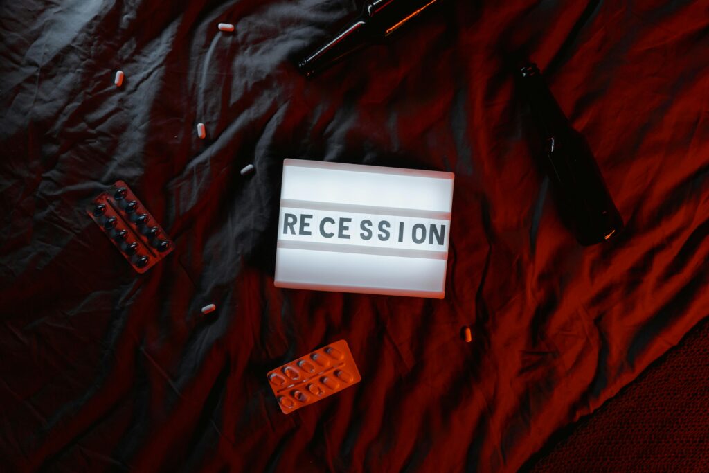 A dimly lit scene with a lightbox displaying the word "RECESSION" on a dark red bedspread. Surrounding the lightbox are scattered pills, blister packs, and empty beer bottles, creating a somber and moody atmosphere.