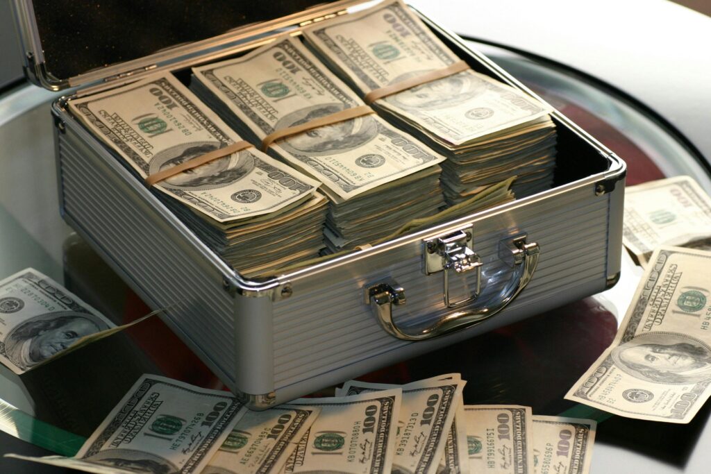 An open silver briefcase filled with stacks of U.S. hundred-dollar bills, neatly bundled with rubber bands, placed on a glass table. Additional loose banknotes are scattered around the case.